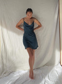 A woman stands barefoot against a white backdrop, looking down with her hands behind her back, wearing the Karima Cowl Neck Slip Dress - Mini Indigo by Rimmba | Sustainable Clothing. Soft natural light casts gentle shadows on the scene.