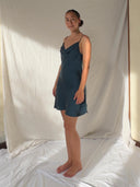 A woman stands barefoot on a white sheet, wearing the Rimmba Karima Cowl Neck Slip Dress - Mini Indigo. She faces slightly to the side with a neutral expression against a backdrop of light, draped fabric.