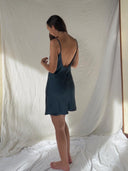 A woman with long brown hair stands barefoot on a white sheet, wearing the Karima Cowl Neck Slip Dress - Mini Indigo by Rimmba | Sustainable Clothing. She faces away toward a light fabric backdrop in soft natural lighting.