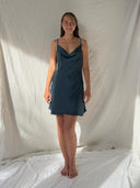 A woman stands barefoot on a white sheet, smiling softly in the Karima Cowl Neck Slip Dress - Mini Indigo by Rimmba | Sustainable Clothing. Her straight brown hair frames her face against a draped, light-filled fabric background.