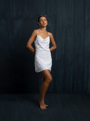 A woman stands barefoot against a dark backdrop, wearing the Karima Dress—an embroidered white cotton mini dress by Rimmba | Sustainable Clothing. With thin straps, she poses serenely, hands behind her back and a pink flower in her hair.