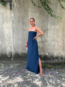 A woman stands barefoot on a paved surface, wearing the Kelly Strapless Silk Dress by Rimmba | Sustainable Clothing. She poses with one hand on her hip before a weathered concrete wall draped with green vines.