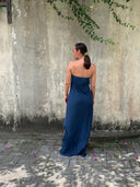 A woman in a strapless indigo Kelly Strapless Silk Dress by Rimmba | Sustainable Clothing stands with her back to the camera on stone pavement, facing a weathered concrete wall beneath hanging green leaves and scattered pink petals.