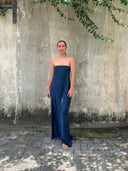 A person stands barefoot on a stone path in front of a weathered wall, wearing the Kelly Strapless Silk Dress by Rimmba | Sustainable Clothing. Green vines hang above, with scattered pink petals on the ground.