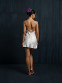 A woman with dark hair in a purple-flower bun stands barefoot, back to the camera, wearing the Rimmba The Brand Lily White Silk Dress - Mini against a dark, textured background.