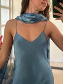 A person wearing the Rimmba Lily Silk Dress - Aquamarine Indigo and a sheer blue scarf is indoors, taking a mirror selfie. The image focus is on their upper body and part of their face.