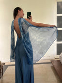 A woman stands indoors taking a mirror selfie, wearing the Lily Silk Dress - Aquamarine Indigo by Rimmba | Sustainable Clothing, showing off its flowing open-back design and sheer, draped silk fabric as she holds part of the dress to the side.