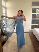 A woman stands barefoot indoors, wearing a Rimmba Lily Silk Dress in Aquamarine Indigo with a matching sheer scarf, taking a mirror selfie in a bright room with large windows and a bed.