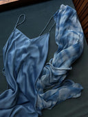 The Lily Silk Dress - Aquamarine Indigo by Rimmba | Sustainable Clothing, featuring thin straps, is displayed on a dark surface with a sheer, blue-and-white tie-dye scarf beside it.