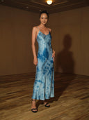 A woman in the Lily Silk Dress - Cosmic Indigo Tiedye by Rimmba | Sustainable Clothing, paired with black heels, stands confidently on a wooden floor in a softly lit room with tan walls, her hair styled in a neat bun.