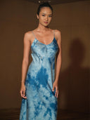 A woman with a neat updo models the Rimmba Lily Silk Dress in Cosmic Indigo Tiedye, standing before a plain brown background and gazing confidently at the camera.
