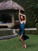 A woman poses gracefully on green grass before a rustic thatched-roof gazebo, wearing the Lily Silk Dress - Indigo by Rimmba | Sustainable Clothing, crafted with natural dye and framed by a lush leafy hedge in the background.