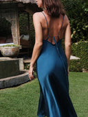 A woman in the Rimmba Lily Silk Dress in Indigo stands on grass near a rustic outdoor structure and lush greenery, facing away from the camera.