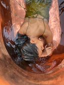 A woman with dark hair relaxes in a copper tub filled with water, wearing the Lily Silk Dress - Mustard Green by Rimmba | Sustainable Clothing. Sunlight shimmers on the surface as she closes her eyes in peaceful repose.