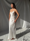 A woman wearing the Lily White Silk Dress by Rimmba The Brand stands barefoot on a white fabric backdrop, softly lit by sunlight. She has long dark hair and gazes to the side with a gentle expression.