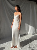 A woman stands barefoot on a white fabric backdrop, wearing the elegant Lily White Silk Dress by Rimmba The Brand. Made from Mulberry Silk with thin straps, she looks serene in soft sunlight with her eyes closed.