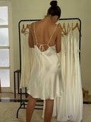 A woman in a silky, open-back Lily White Silk Dress - Mini by Rimmba The Brand stands with her back to the camera, gazing at similar white dresses on a black rack in a softly lit room.