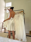 A woman in the Rimmba The Brand Lily White Silk Dress - Mini, backless and ivory, stands by a rack of similar white dresses in a sunlit, minimalist room.