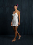 A woman with dark hair and a pink flower stands barefoot against a dark textured background, wearing the Lily White Silk Dress - Mini by Rimmba The Brand, featuring thin straps and a V-neckline.