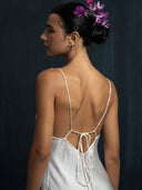 A woman with dark hair in a bun decorated with purple flowers wears the Lily White Silk Dress - Mini by Rimmba The Brand, featuring delicate thin straps and a low back, standing against a dark, textured background and gazing forward.