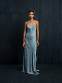 A woman with slicked-back hair and a small purple flower stands against a dark, textured background wearing the Rimmba Lily Silk Dress in Cosmic Indigo Tiedye.