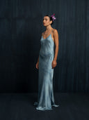 A woman stands before a dark, textured background wearing the Lily Silk Dress - Cosmic Indigo Tiedye by Rimmba | Sustainable Clothing. She has flowers in her hair and gazes calmly to the side.