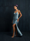 A woman stands barefoot against a dark backdrop in the Lily Silk Dress - Cosmic Indigo Tiedye by Rimmba | Sustainable Clothing, with thin straps, a high slit, flowers in her hair, and strikes a graceful pose while gazing downward.