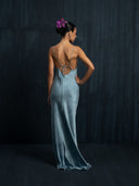 A woman with a purple flower in her hair wears the Lily Silk Dress - Cosmic Indigo Tiedye by Rimmba | Sustainable Clothing, standing against a dark textured background and facing away from the camera.