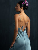 A woman with an elegant updo and purple flowers in her hair wears the Lily Silk Dress – Cosmic Indigo Tiedye by Rimmba | Sustainable Clothing, standing against a dark, textured background and looking downward, turned slightly to the side.