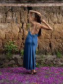 Back-view of long hair woman wearing Rimmba's Lily silk dress midi indigo backless with thin straps. Her face looked to the her arm against a natural wall.