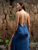 Close-up back view of woman wearing Mulberry silk midi dress indigo backless with thin straps. Her face looked to the side against a natural wall.