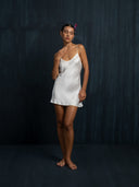 Barefoot woman standing with her eyes closed wearing Mulberry silk mini dress white against a dark background.