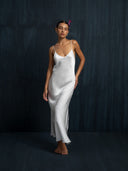A woman stands barefoot against a dark, textured background, wearing a long Lily White Silk Dress by Rimmba The Brand with thin straps. With a flower in her hair and closed eyes, she exudes peaceful elegance.