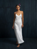 A woman stands barefoot against a dark textured background, wearing the Lily White Silk Dress by Rimmba The Brand. The long white silk dress has thin straps, and she wears her hair slicked back with a purple flower, exuding calm confidence.