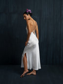 A woman in a Lily White Silk Dress by Rimmba The Brand, featuring thin straps and a side slit, stands barefoot against a dark textured wall, facing sideways with flowers in her hair and her gaze down.