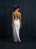 A woman stands barefoot against a dark textured wall wearing the Lily White Silk Dress by Rimmba The Brand—a long, backless white silk gown with thin straps and ties. Her hair is in a bun with purple flowers as she faces away from the camera.