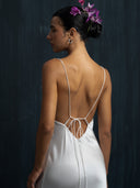 A woman with flowers in her hair wears the Lily White Silk Dress by Rimmba The Brand, featuring thin straps. She stands against a dark background, facing sideways to reveal her back and profile.