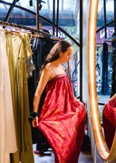 A woman admires herself in front of a mirror, holding out the Parvati Red-Eco Print Dress by Rimmba | Sustainable Clothing. Around her, racks display more dresses featuring similar eco-friendly red prints.