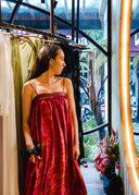 A woman in the Parvati Red-Eco Print Dress by Rimmba | Sustainable Clothing stands by a clothing rack, gazing at a curved mirror in a boutique filled with plants and views of the city street through the windows.