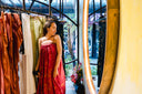 A woman in a red dress stands among clothing racks in a boutique, looking at herself in a large mirror with a light wooden frame. The shop has large windows and a floral arrangement on the floor.