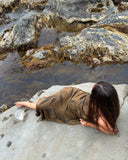 Karunia Fischer's lying on a smooth rock surface near a rocky shoreline. The person appears to be partially covered in wet sand, blending with the natural surroundings. They are positioned on their side, propped up slightly on one arm, with their legs extended.