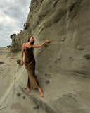 Karunia Fischer's is standing barefoot on a narrow sandy ledge beside a rugged cliff by the beach. She is wearing a long, flowing brown slip dress and appears relaxed as she lightly touches the textured rock wall with one hand.