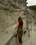 A woman is walking barefoot along a narrow ledge carved into a sandy, textured cliff. The photo is taken from behind, showing her long, wavy brown hair flowing down her back. She is wearing a fitted, earthy-toned slip dress that moves naturally with her steps.