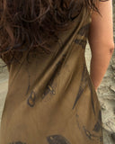 A close-up view shows the back of a woman wearing a silky, earth-toned slip dress. The fabric has a smooth, slightly reflective texture and features abstract, organic patterns in darker shades, resembling natural stains or brush-like markings