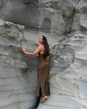 Karunia Fischer standing barefoot against a textured rock formation cliff. She is wearing a long, brown dress and has long, dark hair flowing down her back.