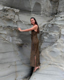 a woman standing barefoot against a large, textured rock formation. She is wearing a long, sleeveless brown dress with a slit on the side, and her long dark hair falls naturally over her back. She is turned slightly toward the camera, smiling, while placing one hand on the rock surface as if feeling its texture.