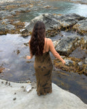 a woman standing on a smooth rock surface near the edge of a shallow coastal area. She is barefoot and wearing a long brown dress with a subtle leaf-like pattern. Her long, wavy dark hair falls down her back as she faces away from the camera, looking out toward the water.
