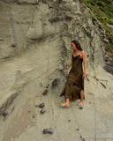 Karunia Fischer's walking barefoot along a sloped, sandy rock surface near a cliff. she's wearing a brown dress that blends with the earthy tones of the surroundings. Their posture is relaxed, and they appear to be looking slightly downward as they step carefully over scattered small stones.