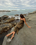 Karunia Fischer's sitting on a large, smooth rock by the ocean. They are wearing a brown dress and leaning back with one hand resting on the rock for support. Their legs are stretched out in front of them, with bare feet placed on the stone surface.