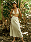 A woman stands barefoot outdoors before large tropical leaves, wearing Rimmba The Brand’s Meeko - Burgundy Maxi Silk Skirt with a matching top and headband.